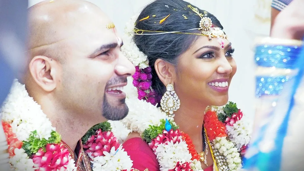 A bride in a red lehenga with gold embroidery alongside a groom in a cream sherwani